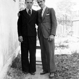 Two men in suits standing outdoors near a wall.