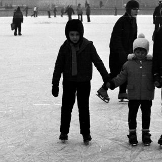 Two children ice skating with others in a snowy park.