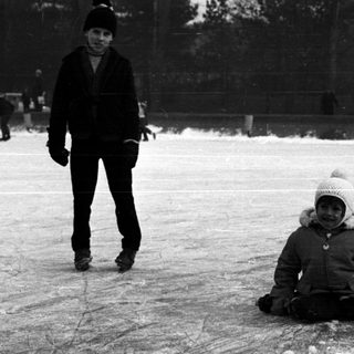 Child sits on ice rink; another skates nearby.