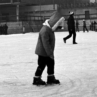 People ice skating outdoors near a building and trees.