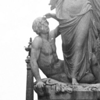 Here's a brief summary of the image: Person sits on a bench near a statue in a garden.