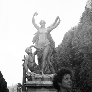 Here's a brief summary of the image: Person sits on a bench near a statue in a garden.