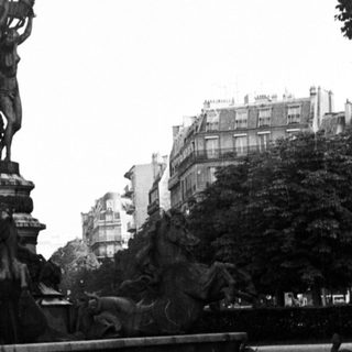 Monumental fountain with statue, reflecting in water, cityscape backdrop.