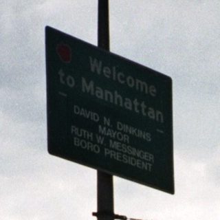 Man stands near Welcome to Manhattan sign, towers visible.