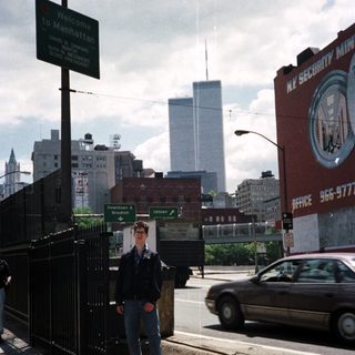 Man stands near Welcome to Manhattan sign, towers visible.