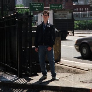 Man stands near Welcome to Manhattan sign, towers visible.
