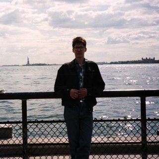Man stands near water with Statue of Liberty in background.