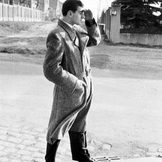Man in coat stands outdoors, shading his eyes.