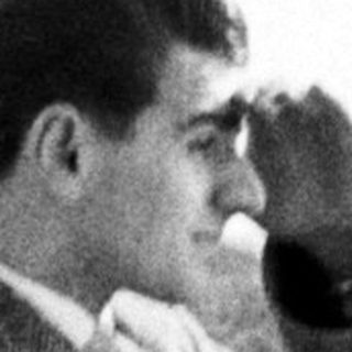 Man in coat stands outdoors, shading his eyes.