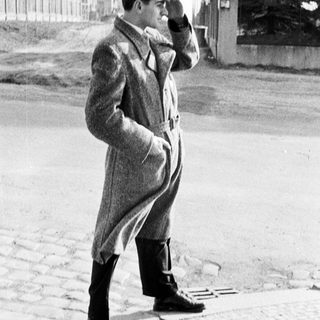 Man in coat stands outdoors, shading his eyes.