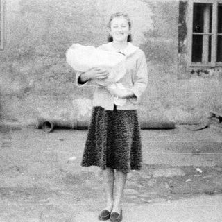 Here's a brief summary of the image: Woman stands holding a baby outdoors, possibly in a yard.