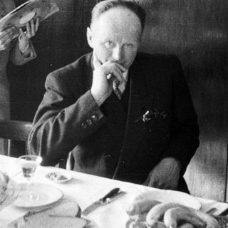 Man at table, boy stands nearby reading.