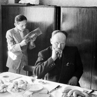 Man at table, boy stands nearby reading.