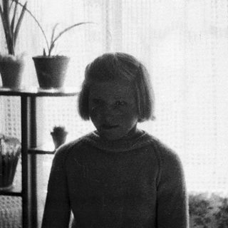 Young girl stands near plants and a window.