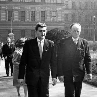 Two men in suits walking outdoors, with others in background.