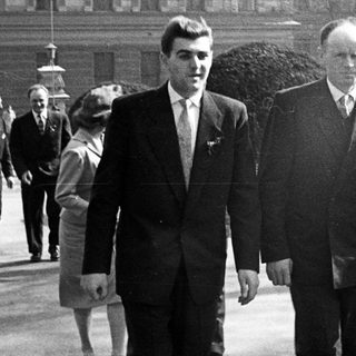 Two men in suits walking outdoors, with others in background.
