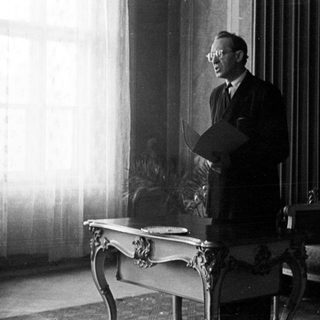 Man stands holding papers in a room with furniture.