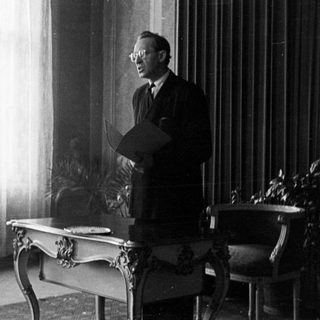 Man stands holding papers in a room with furniture.
