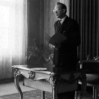 Man stands holding papers in a room with furniture.