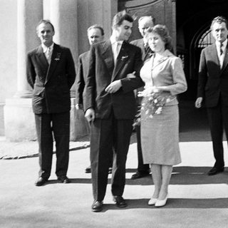 Couple exits building, surrounded by men, in vintage attire.