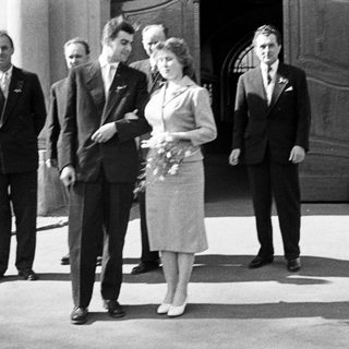 Couple exits building, surrounded by men, in vintage attire.