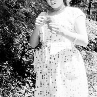 Woman in dress stands outdoors, holding something in her hands.