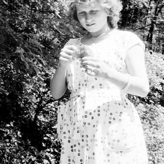 Woman in dress stands outdoors, holding something in her hands.