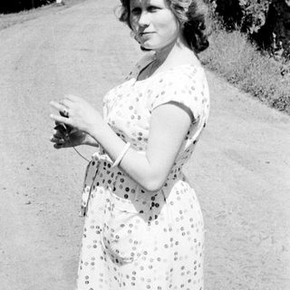 Woman in dress stands on a road, looking at camera.