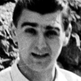 Man stands barefoot in trousers and shirt outdoors.