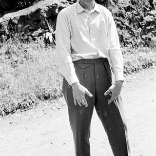 Man stands barefoot in trousers and shirt outdoors.