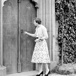 Woman approaches ornate door with ivy-covered wall.