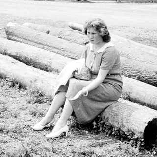 Woman sits on logs, a rooster nearby in the yard.