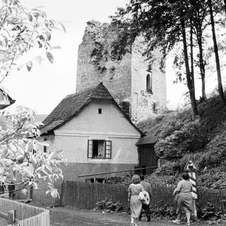 People walk past a ruined tower and a house.