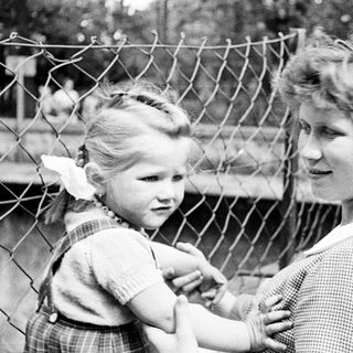 Here's a brief summary of the image: Child and woman near a fence, soldiers in background.