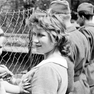 Here's a brief summary of the image: Child and woman near a fence, soldiers in background.