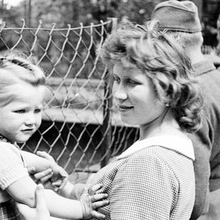 Here's a brief summary of the image: Child and woman near a fence, soldiers in background.