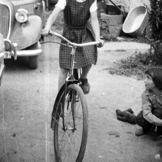 Here's a brief summary of the image: Young girl rides a bicycle near a vintage car.