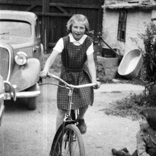 Here's a brief summary of the image: Young girl rides a bicycle near a vintage car.