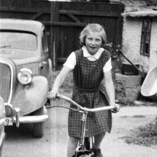 Here's a brief summary of the image: Young girl rides a bicycle near a vintage car.