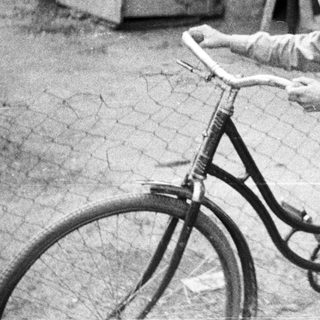 Boy stands near a bicycle, a woman in background.