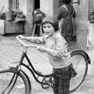 Boy stands near a bicycle, a woman in background.