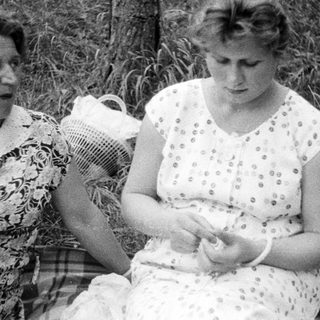 Here's a brief summary of the image: Woman and girl seated outdoors, possibly on a picnic blanket.