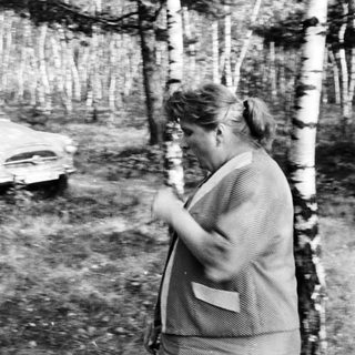Woman stands near a vintage car in a wooded area.