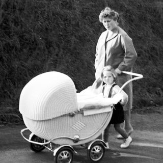 Woman pushes a wicker baby carriage along a road.