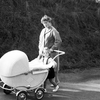Woman pushes a wicker baby carriage along a road.