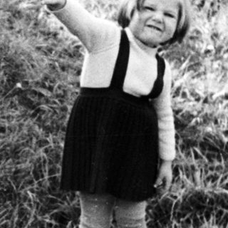 Here's a brief summary of the image: Child stands near a sign in a grassy, rural setting.