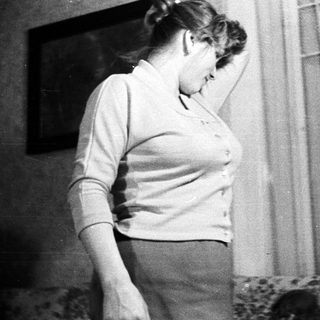 Woman stands near a window, appearing distressed or uncomfortable.