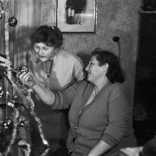 Two women decorate a Christmas tree in a vintage setting.