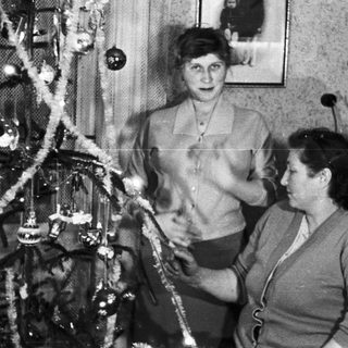 Here's a brief summary of the image: Two women decorate a Christmas tree in a vintage setting.