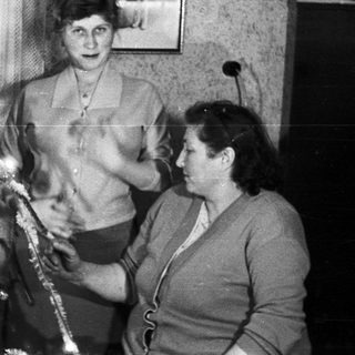 Here's a brief summary of the image: Two women decorate a Christmas tree in a vintage setting.